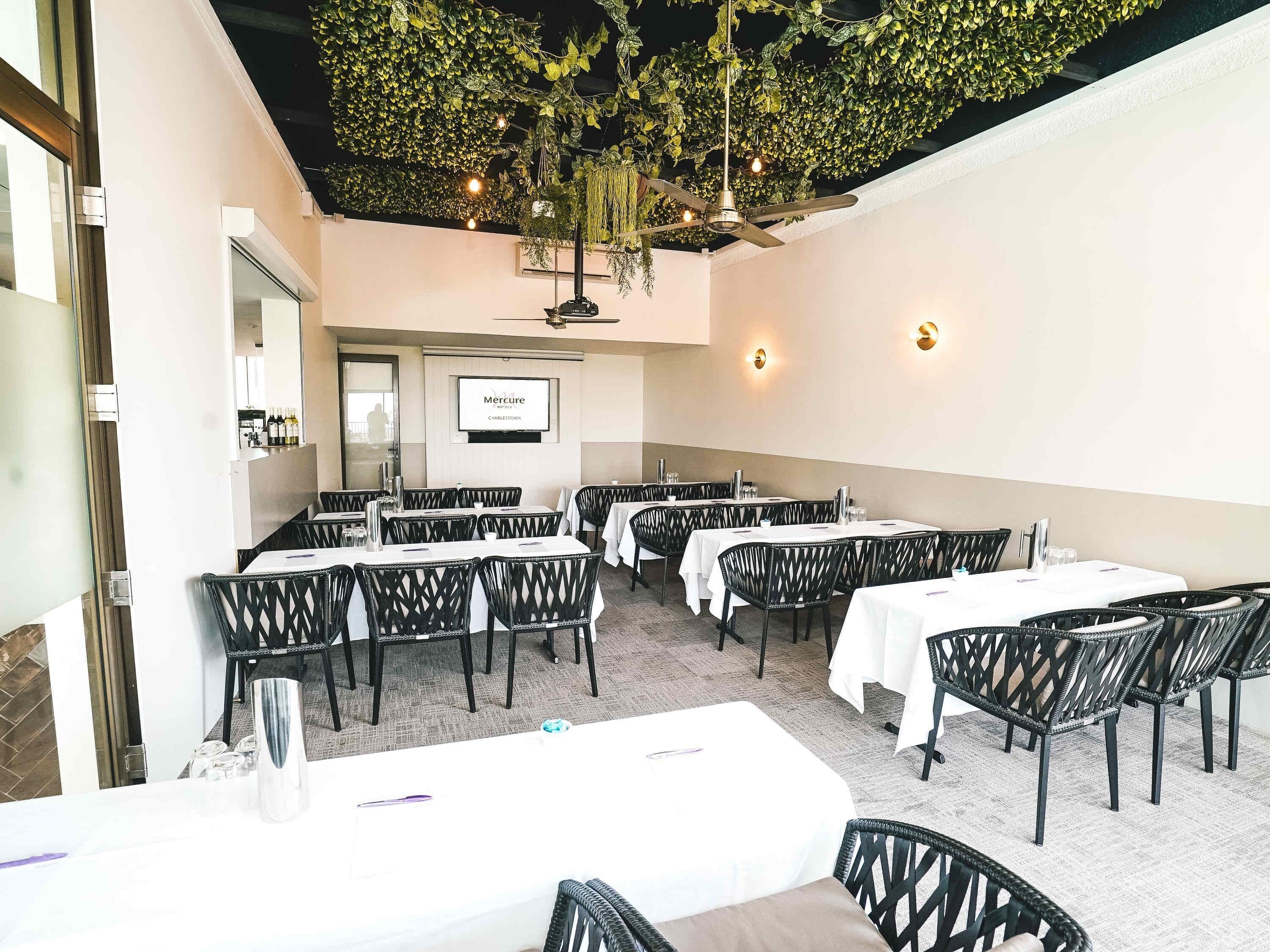 Un restaurante con mesas redondas, sillas de mimbre negro, un jardín en el techo y un letrero colgado en la pared.