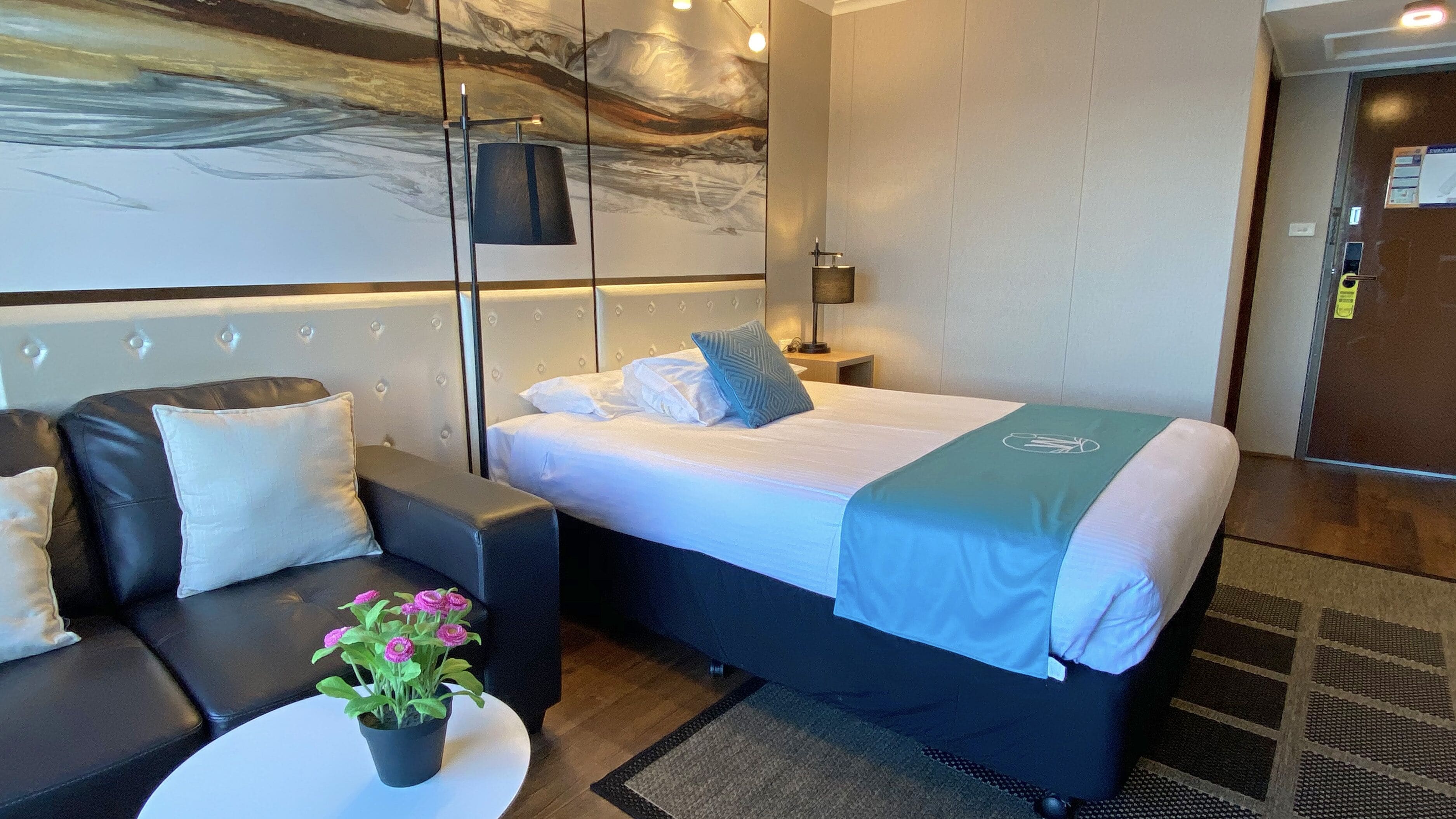 A modern hotel room with a large bed, a sofa, a coffee table with a potted plant, and a wall with a painting.