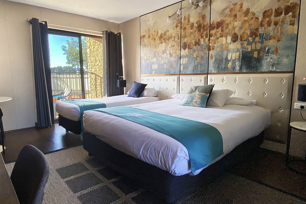 A hotel room with two beds, a desk, and a chair. There is a large window with curtains, a painting on the wall, and a small table with a plant.