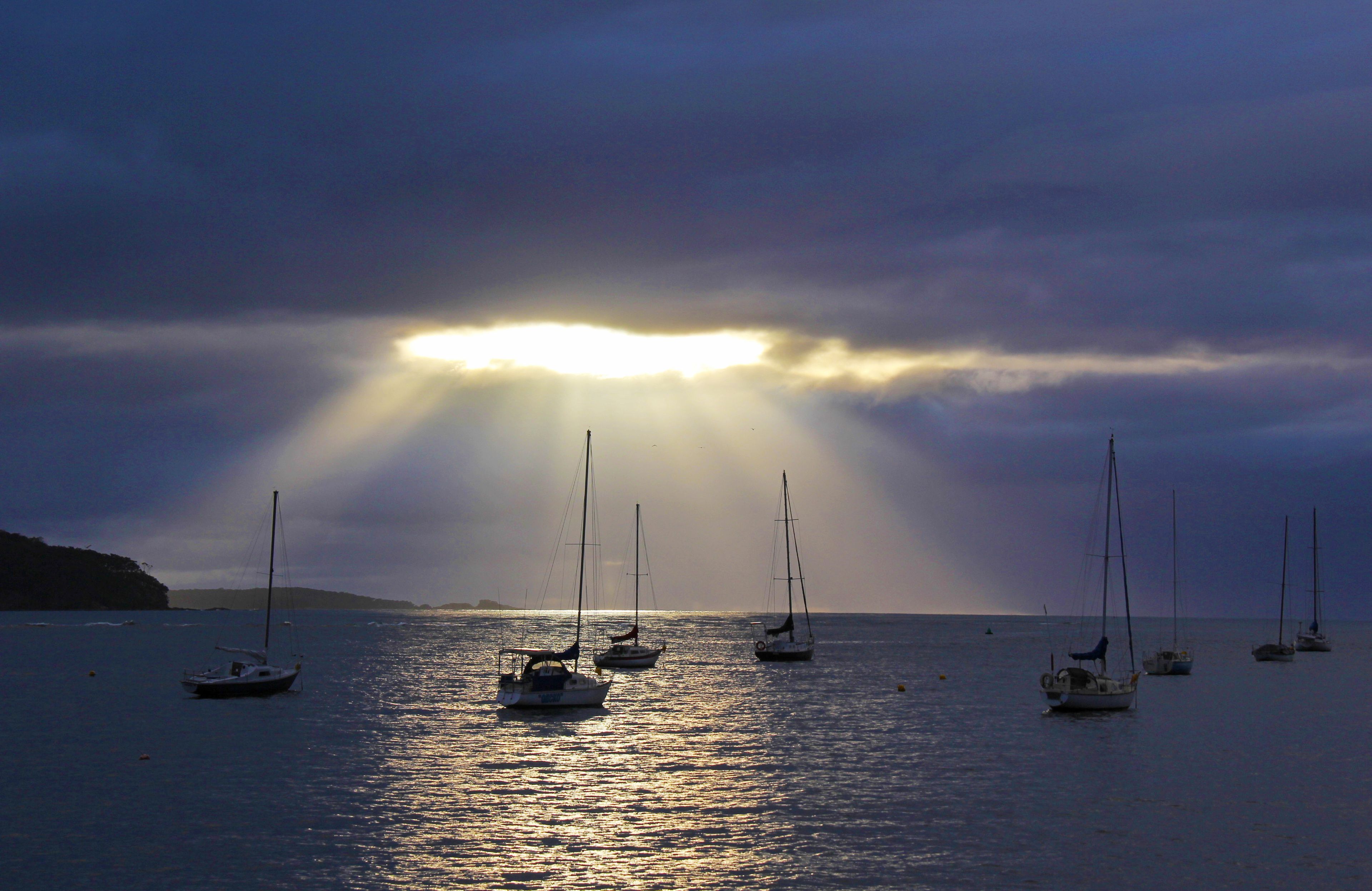 Mar tranquila con varios veleros anclados, bajo un cielo nublado con rayos de sol.