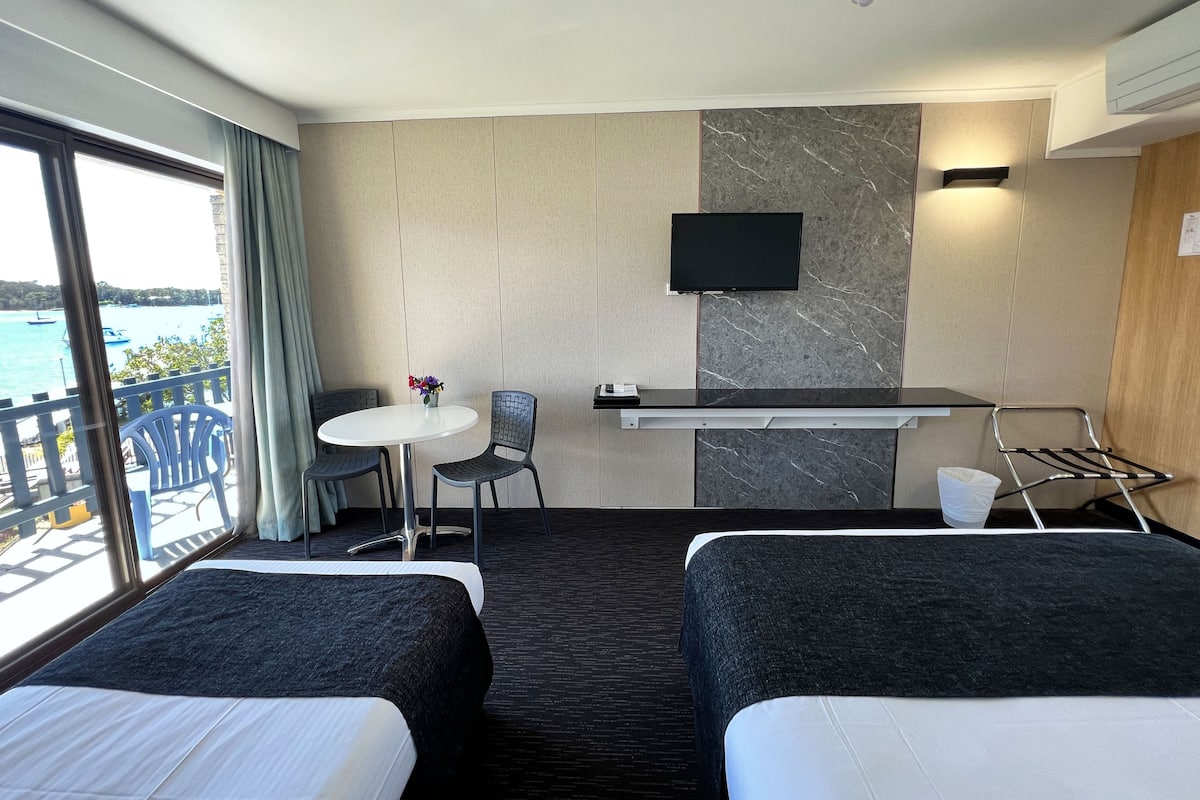 A hotel room with two beds, a small table with chairs, a TV, and a view of the sea.