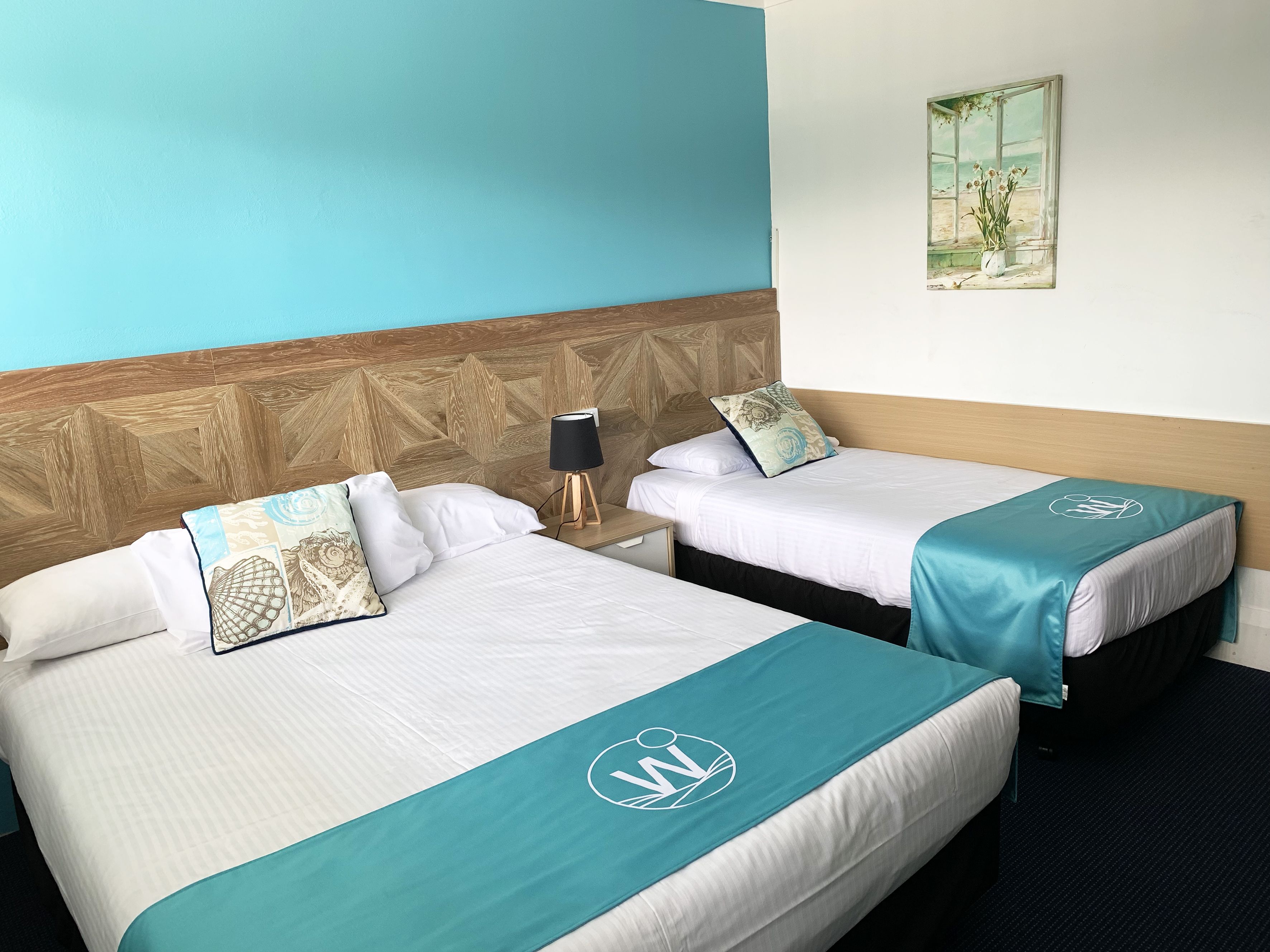 A hotel room with two beds, each with a teal bedspread and a logo, a wooden headboard, a nightstand with a lamp, and a painting on the wall.