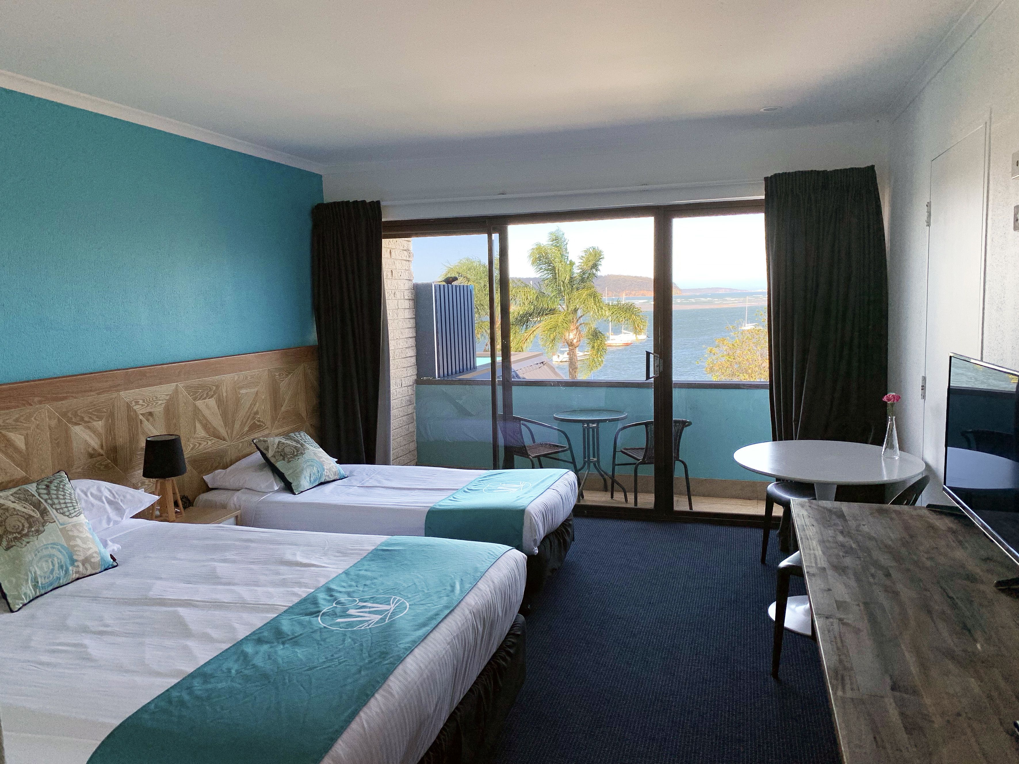 Habitación de hotel con dos camas, un balcón con vistas al mar, una pequeña mesa y un televisor.