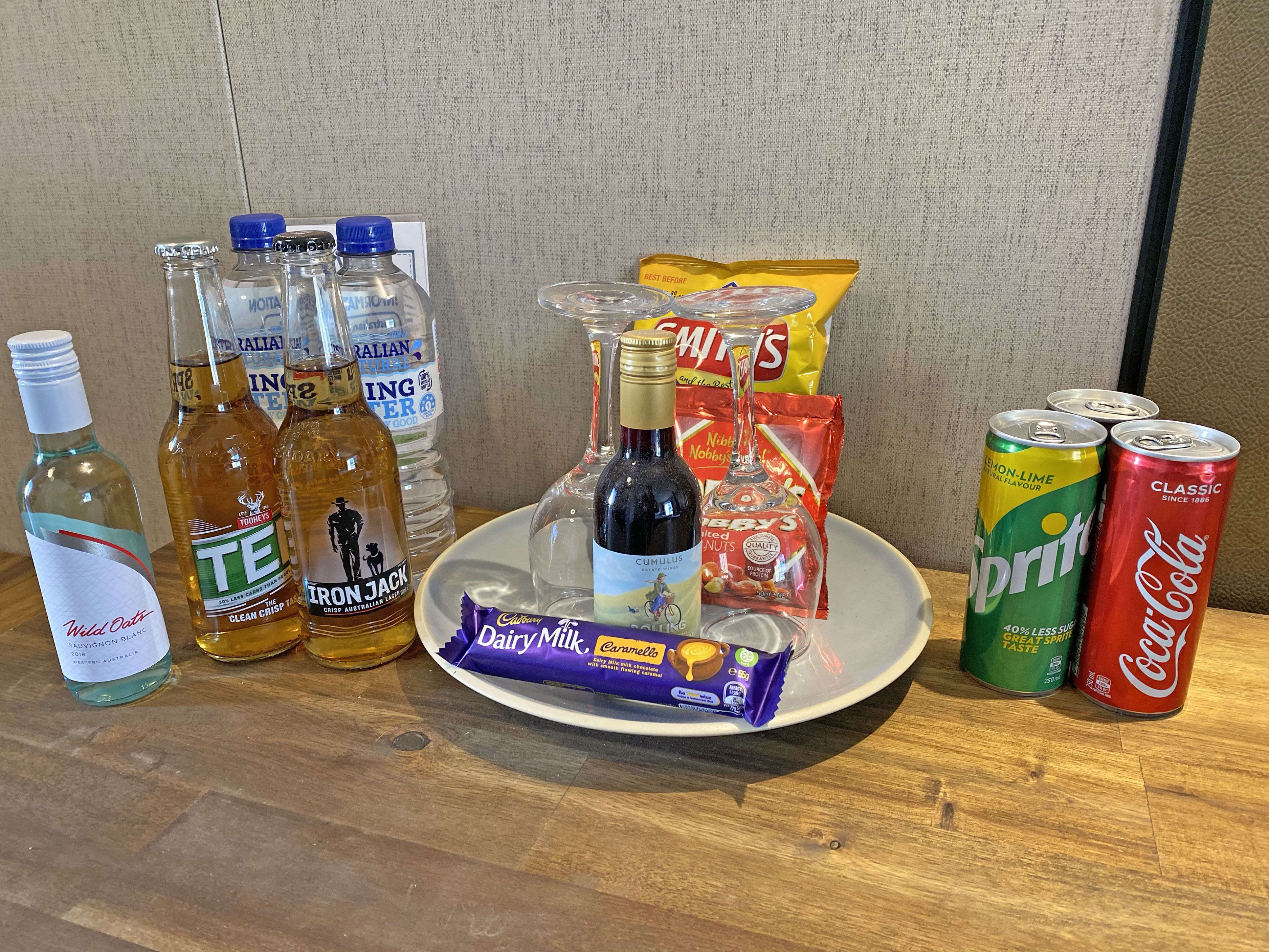 A table with various beverages and snacks including bottles of water, a bottle of wine, cans of soda, and a plate with a chocolate bar and chips.