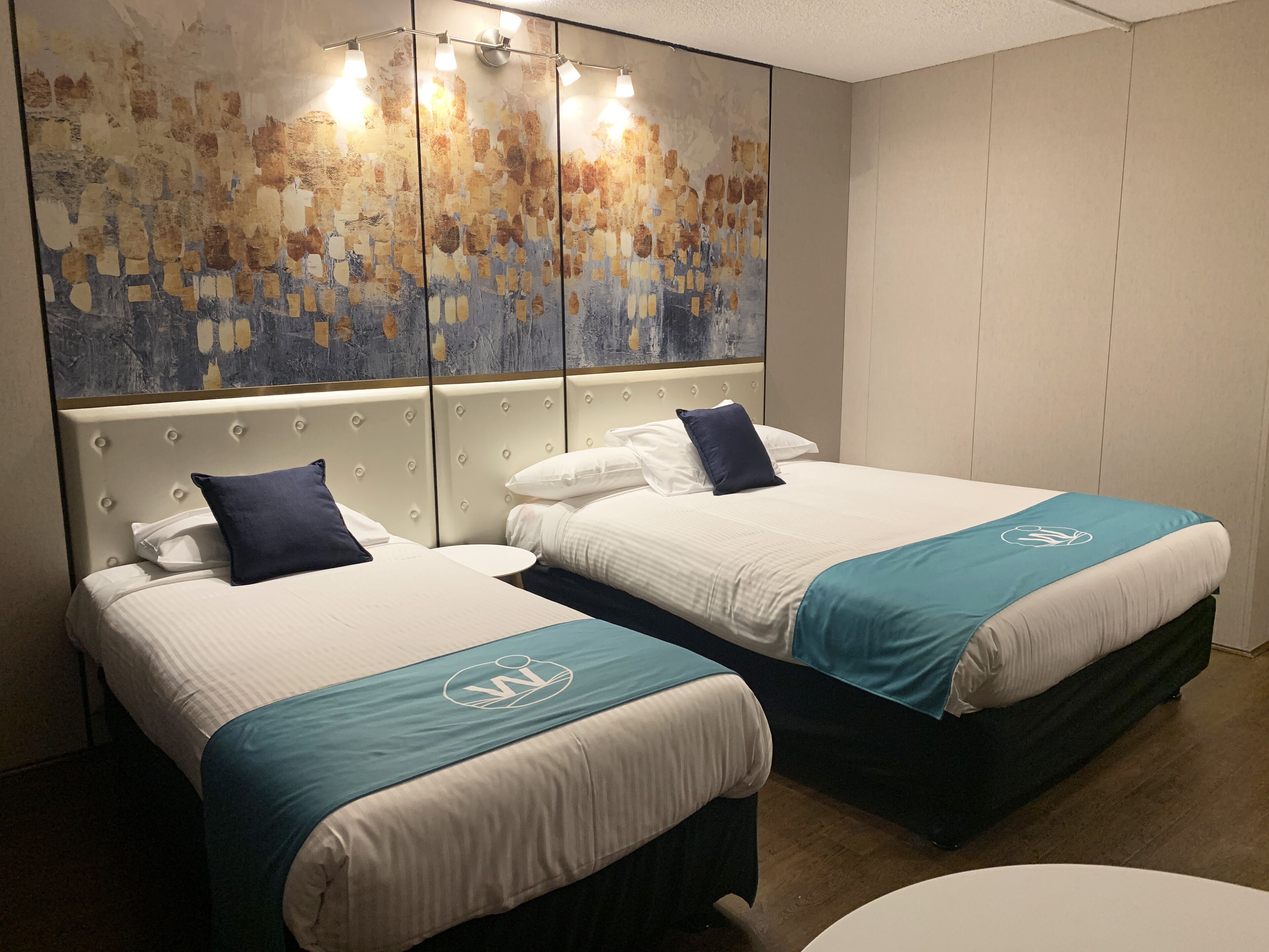 Une chambre d’hôtel avec deux lits, chacun recouvert d’une courtepointe turquoise ornée d’un logo, une grande peinture abstraite au mur et une table ronde au centre.