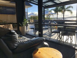 A modern lounge area with a leather sofa, a round wooden table, and a large window overlooking a patio with outdoor seating and umbrellas.