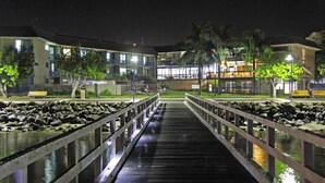 A wooden bridge with railings, leading to a building with multiple balconies and windows, surrounded by trees and a fence.