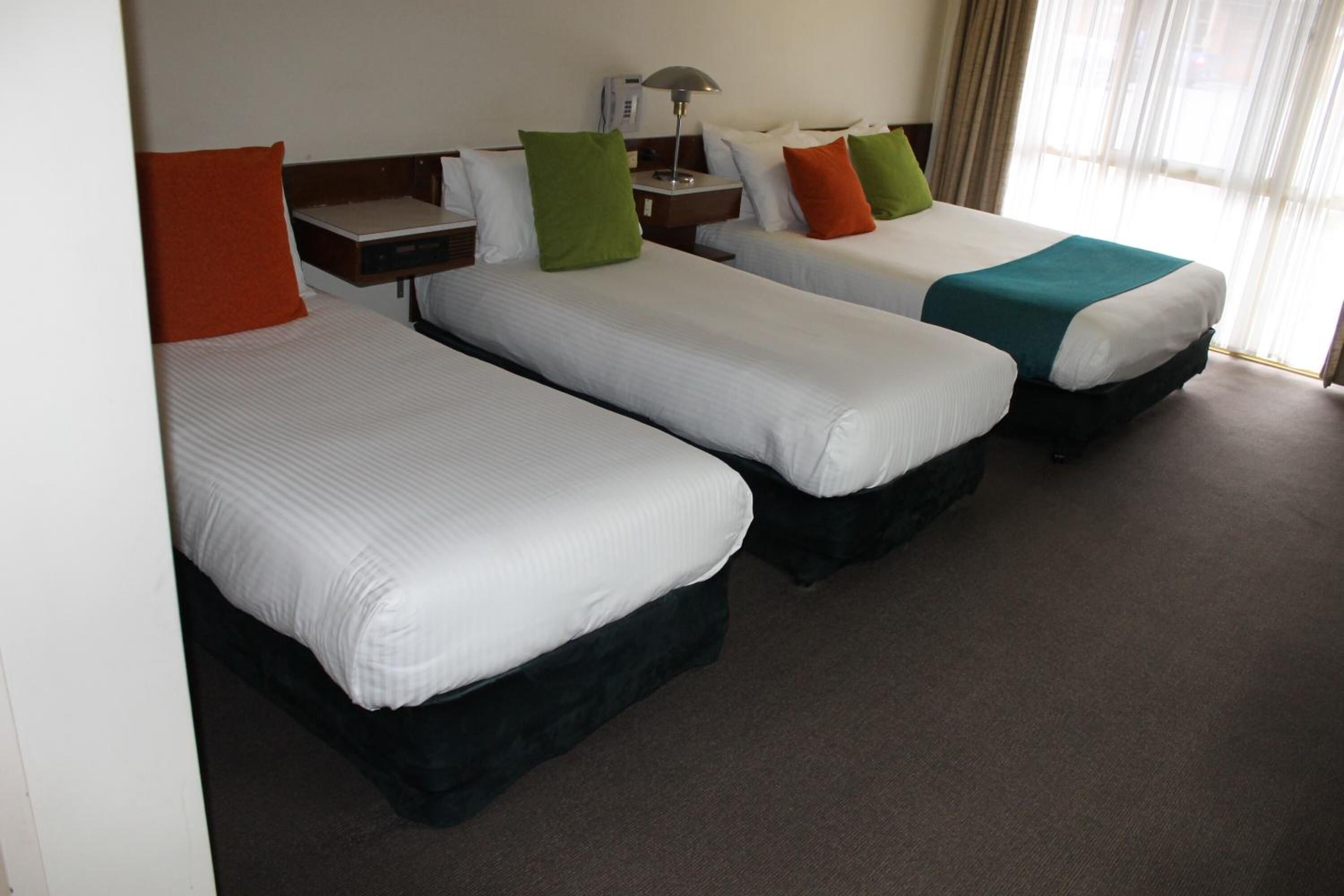 Habitación de hotel con tres camas individuales, cada una con un almohadón de diferente color, una mesita de noche con lámpara, y vista a la calle por la ventana.