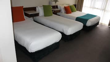 Habitación de hotel con tres camas individuales, cada una con un almohadón de diferente color, una mesita de noche con lámpara, y vista a la calle por la ventana.