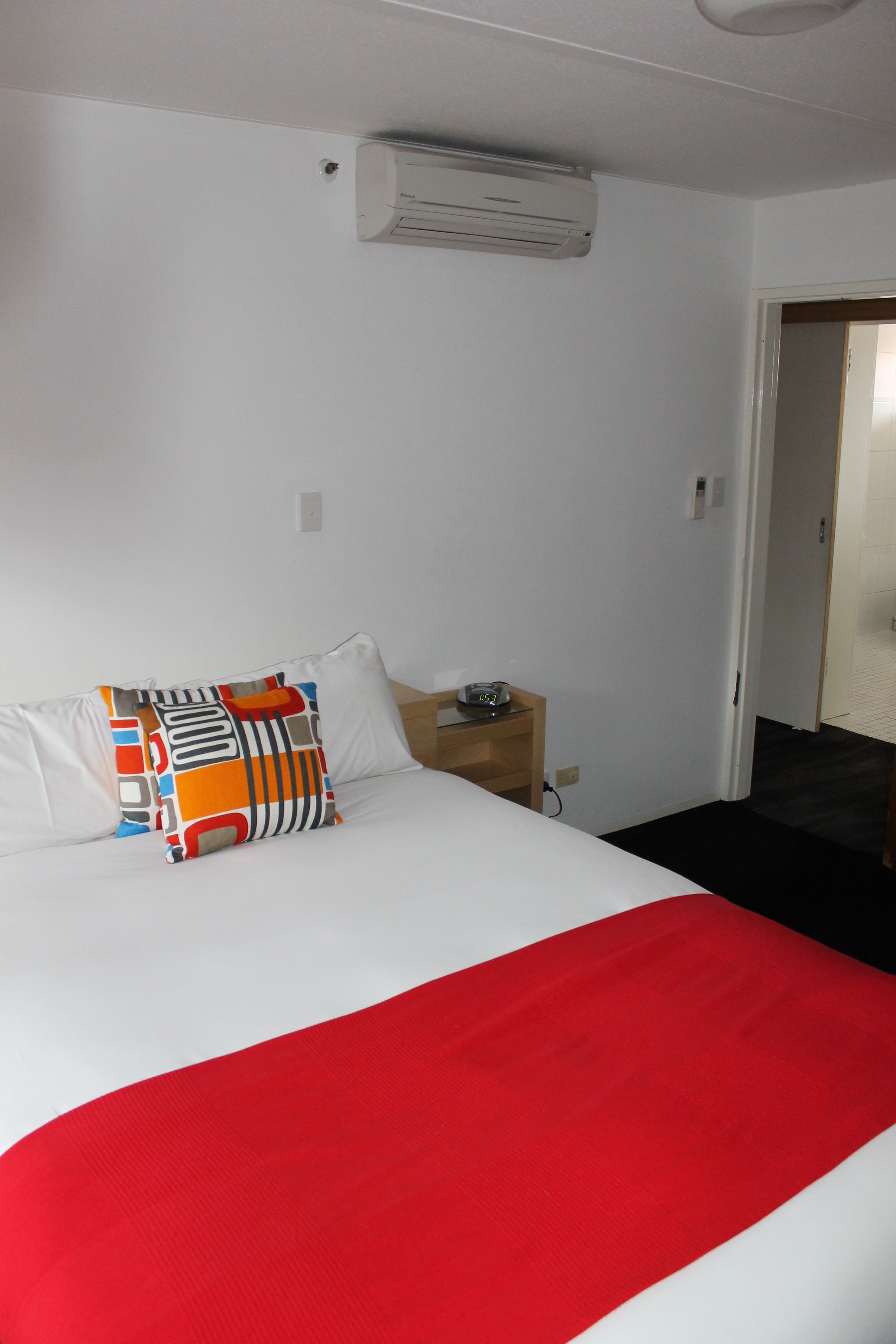 A hotel room with a bed, a nightstand, an air conditioning unit, and a door leading to another room.