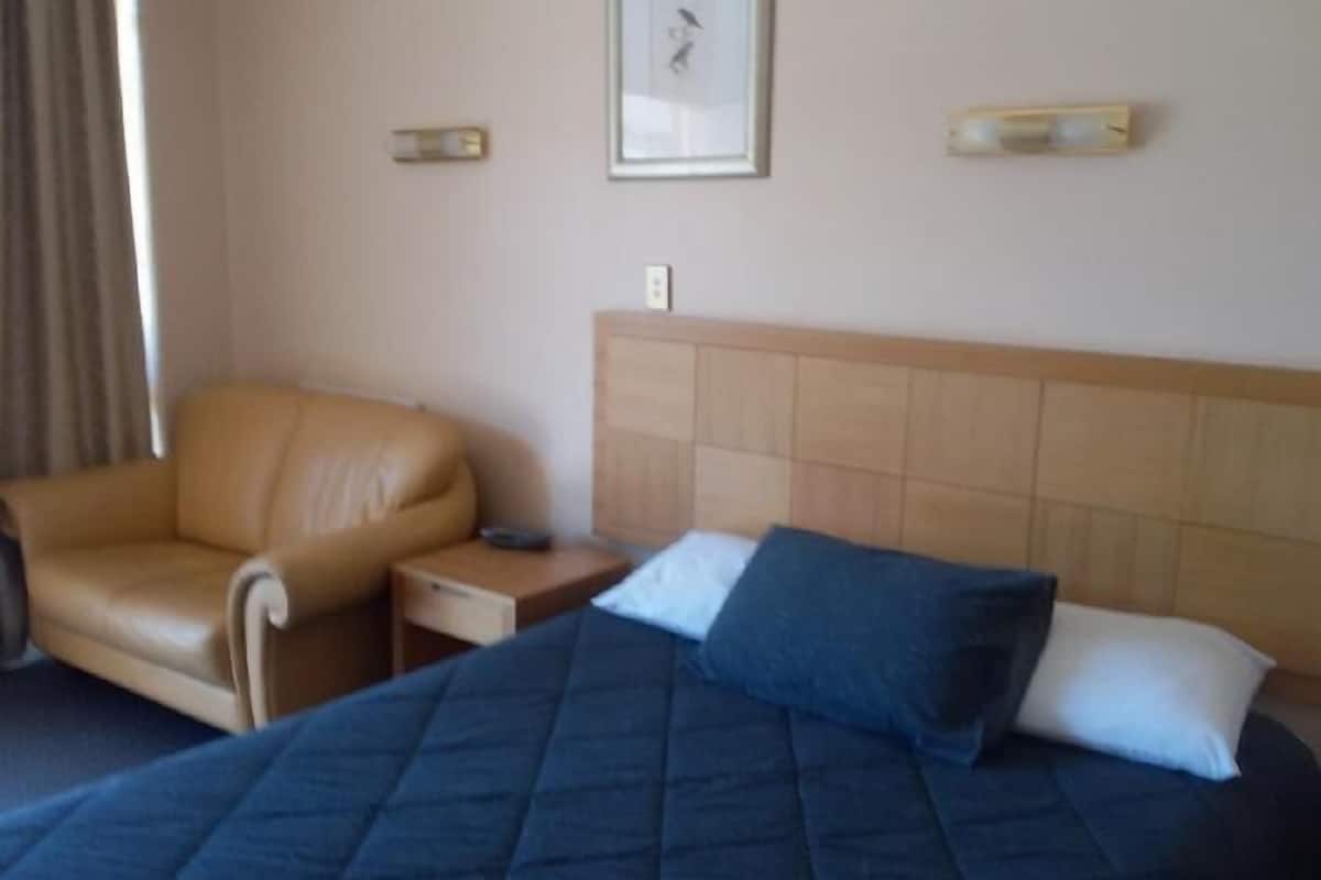 Quarto de hotel com uma cama, uma cadeira, mesa de cabeceira e luminária na parede.