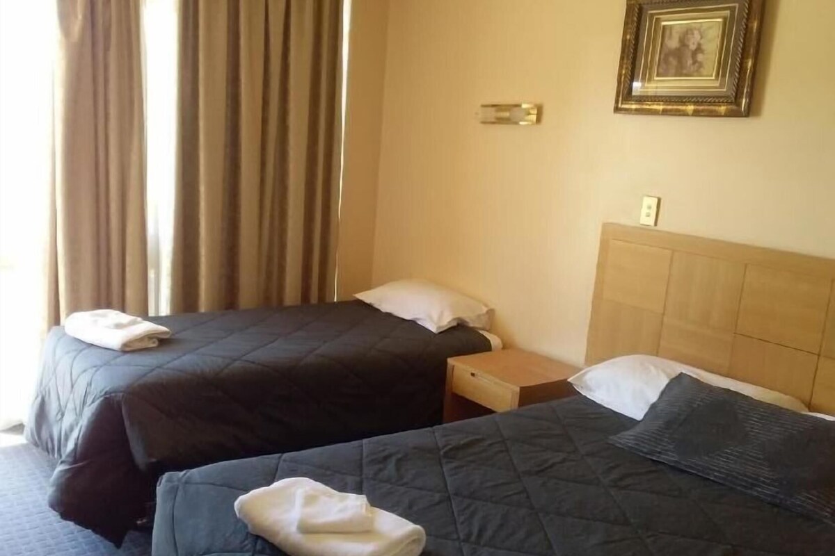Habitación de hotel con dos camas, cada una con una almohada y una toalla doblada. Hay una mesita de noche entre las camas y un marco de fotos en la pared.