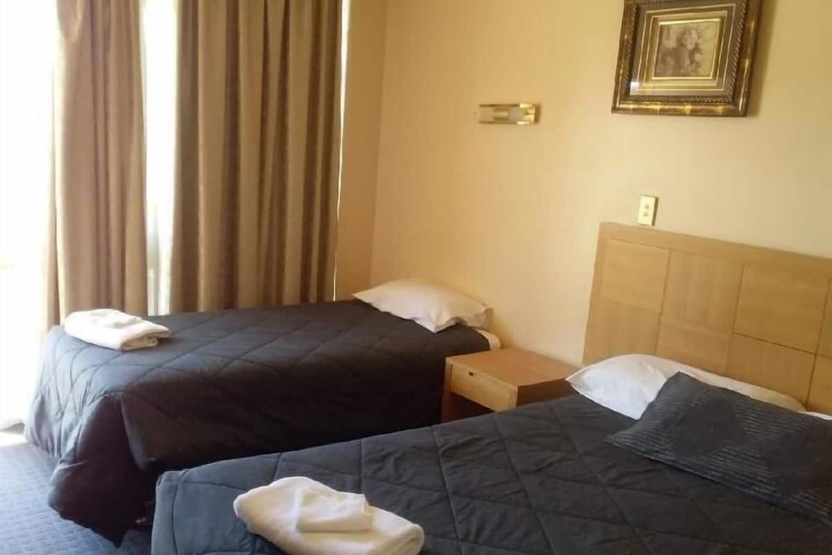 Quarto de hotel com duas camas, cada uma com travesseiro e toalha dobrada. Há uma mesinha de cabeceira entre as camas e um porta-retrato na parede.
