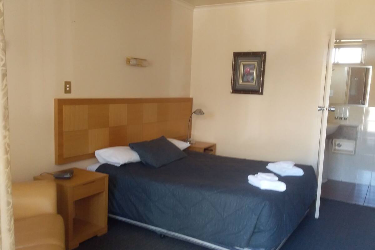 Quarto de hotel com uma cama, mesa de cabeceira, uma cadeira, um abajur, um quadro na parede e um banheiro visível por uma porta aberta.