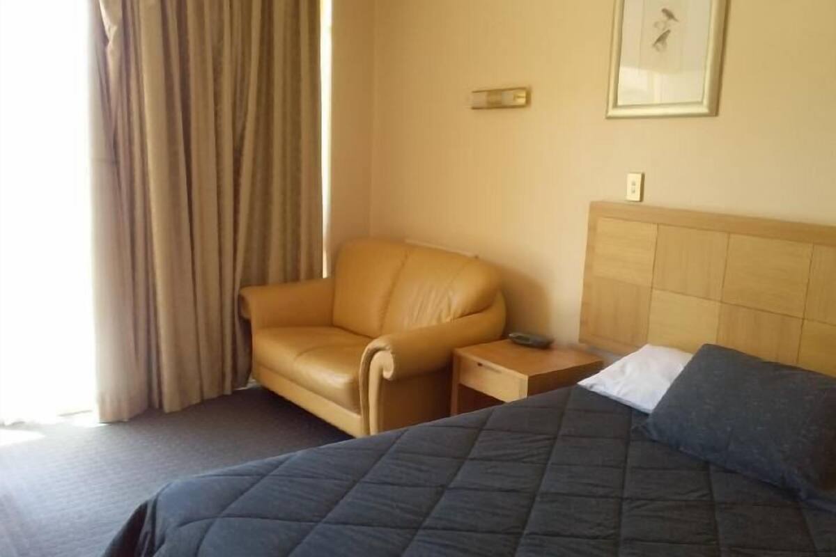 Quarto de hotel com cama, cadeira, mesa de cabeceira e interruptor de luz fixado na parede.