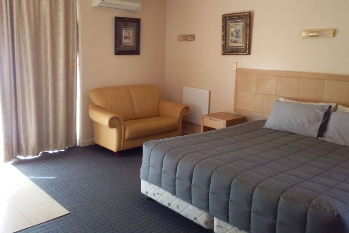 Quarto de hotel com uma cama, uma cadeira, uma mesinha de cabeceira e duas imagens emolduradas na parede.