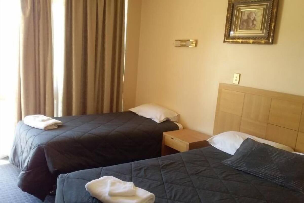 Quarto de hotel com duas camas, cada uma com colcha acolchoada escura e travesseiro branco. Há uma cabeceira de madeira no lado direito e uma pequena mesa de cabeceira de madeira com gaveta. Uma moldura com um quadro está pendurada na parede acima da cabeceira.