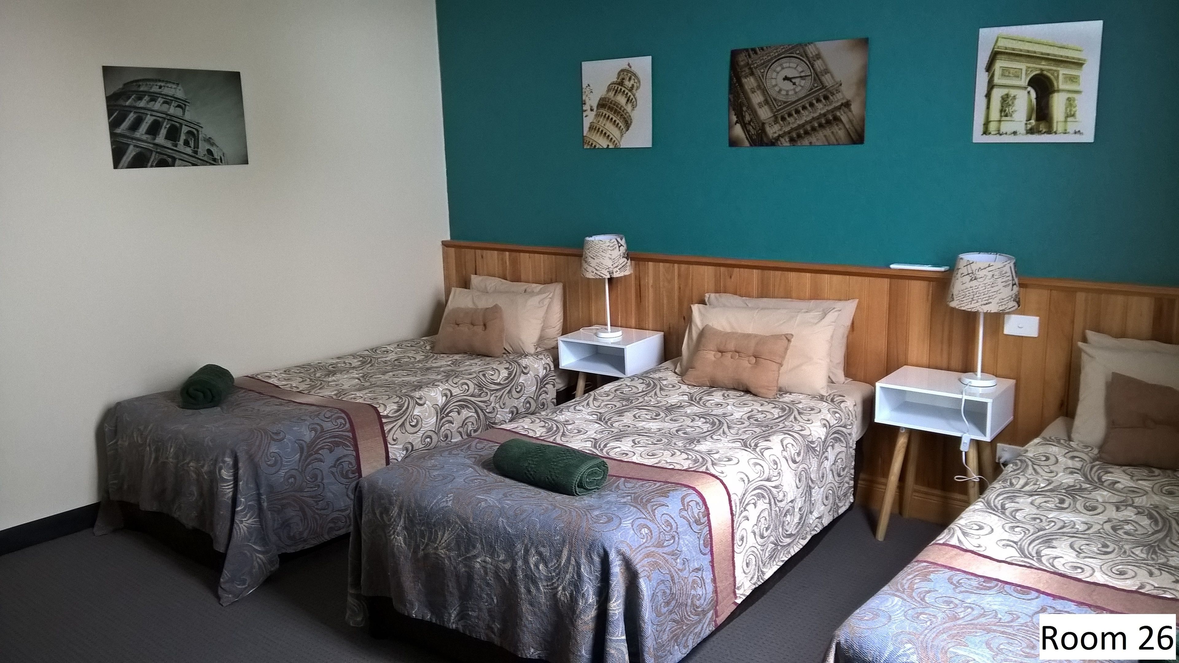 Una habitación de hotel con tres camas, cada una con ropa de cama y almohadas estampadas. Hay lámparas de pared sobre mesitas de noche y un reloj en la pared.