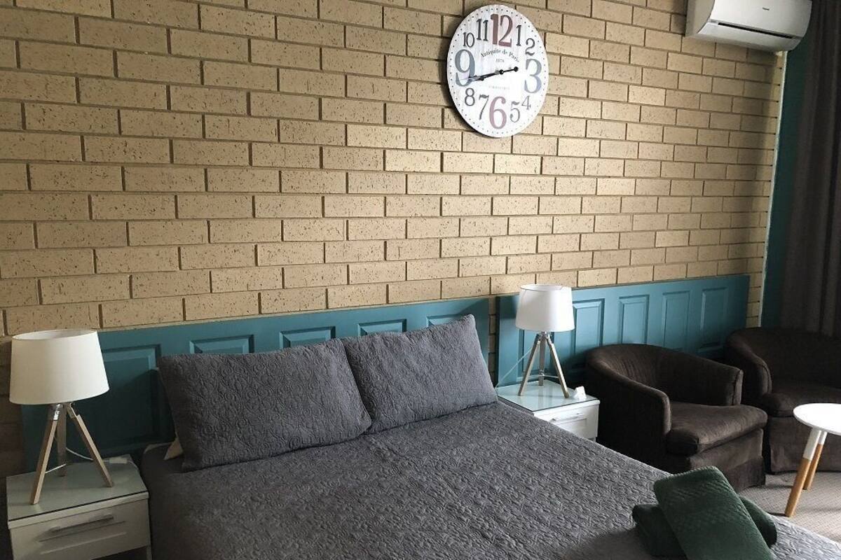 Une chambre avec un mur en briques, une horloge, un lit, deux tables de chevet, une lampe, une chaise et une petite table.