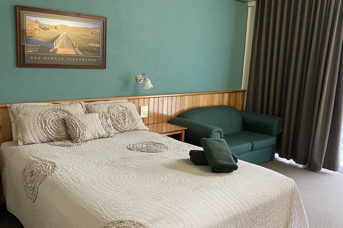 Ett hotellrum med en säng, en grön fåtölj, en inramad tavla på väggen och ett fönster med gardiner.