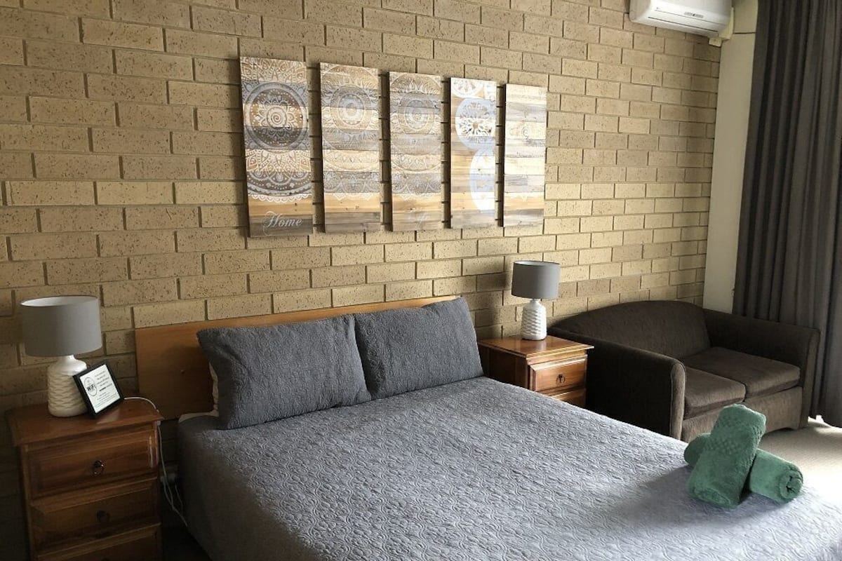 Une chambre d’hôtel avec un mur en briques, un lit recouvert d’une couette grise, deux tables de chevet avec des lampes, une chaise et une œuvre d’art accrochée au mur.