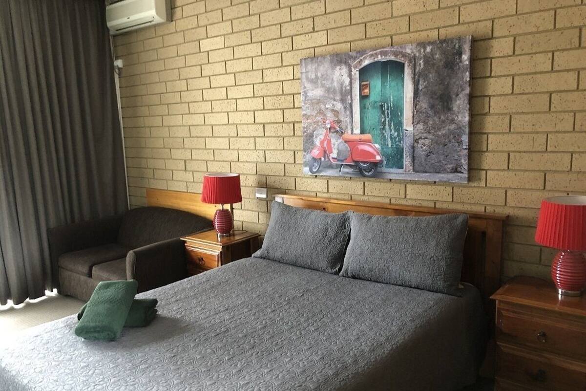 Ett hotellrum med en säng, en stol, ett nattduksbord, en lampa och en tavla med en moped på väggen.