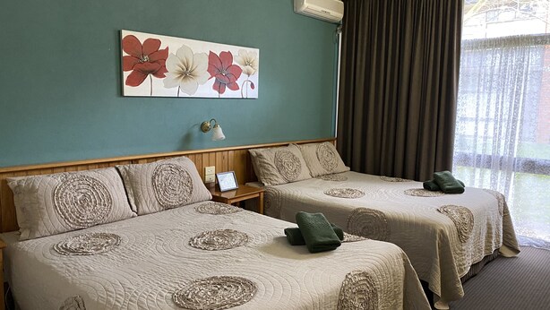 Dos camas con cabeceras a juego y almohadas decorativas, una mesita de noche con lámpara, y un cuadro floral en la pared.