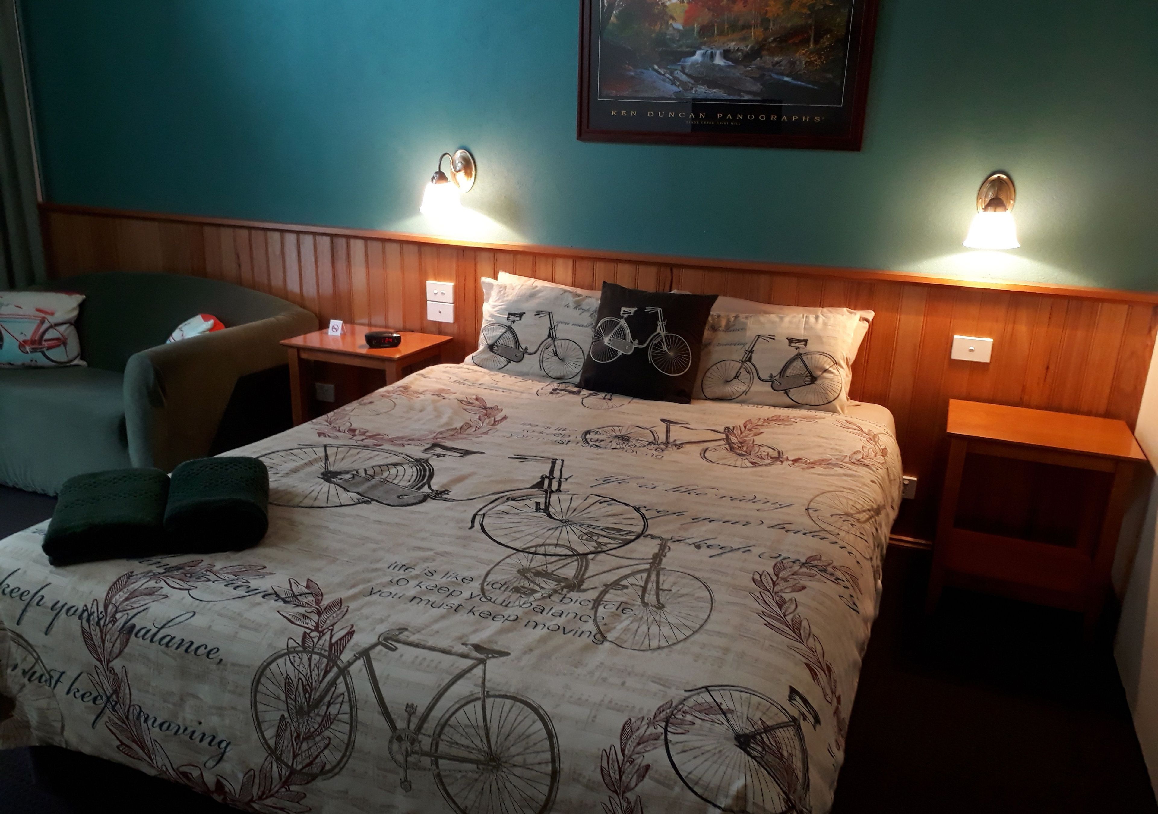 A hotel room with a bed featuring a bicycle-themed bedspread, two bedside lamps, a chair, and a framed picture on the wall.