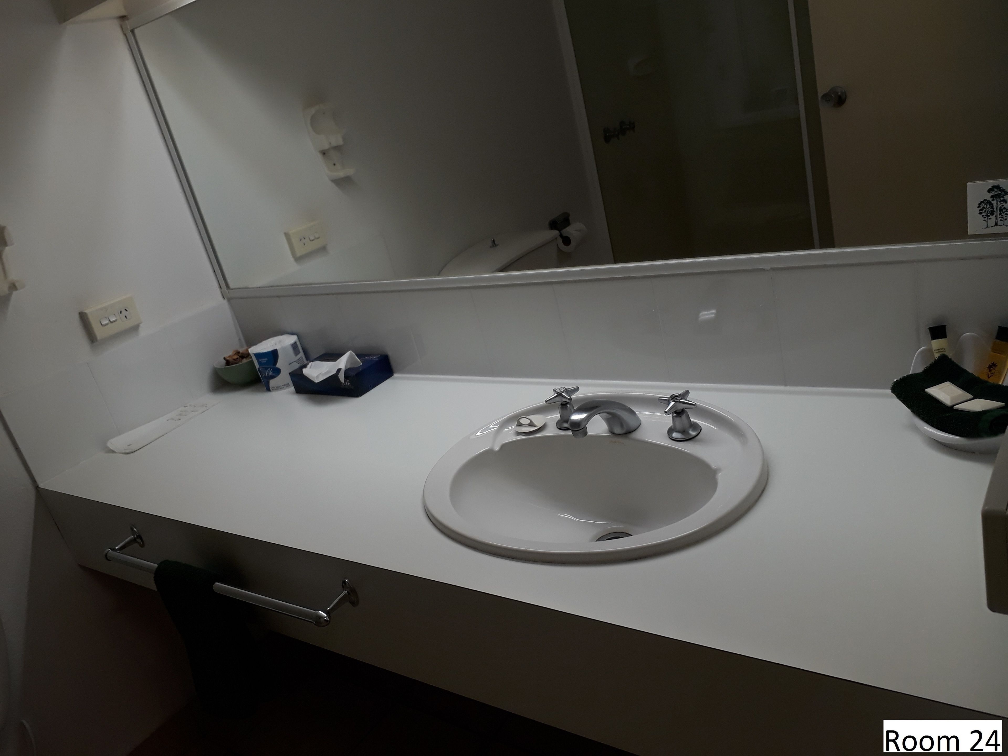 A bathroom with a white sink, two faucets, a mirror, a towel rack, and a toilet. There are toiletries on the counter.