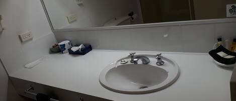 A bathroom with a white sink, two faucets, a mirror, a towel rack, and a toilet. There are toiletries on the counter.