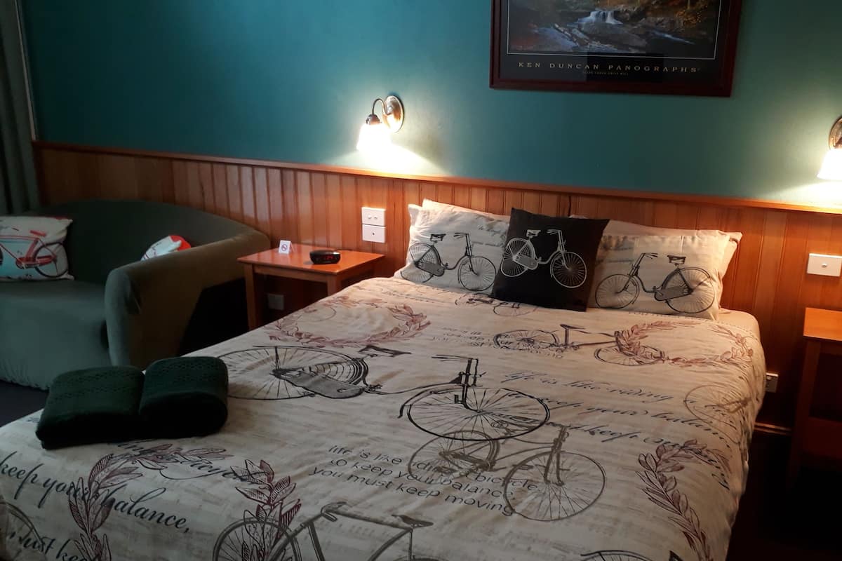 Ett välordnat hotellrum med en säng som har ett sängöverkast med cykeltema, två sänglampor, ett litet bord med en klocka och en stol med en kudde.
