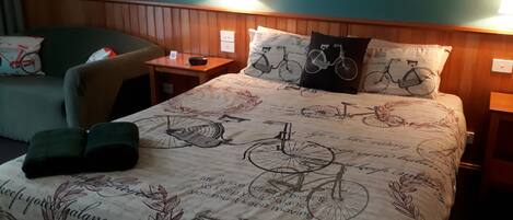 A neatly arranged hotel room with a bed featuring a bicycle-themed bedspread, two bedside lamps, a small table with a clock, and a chair with a cushion.