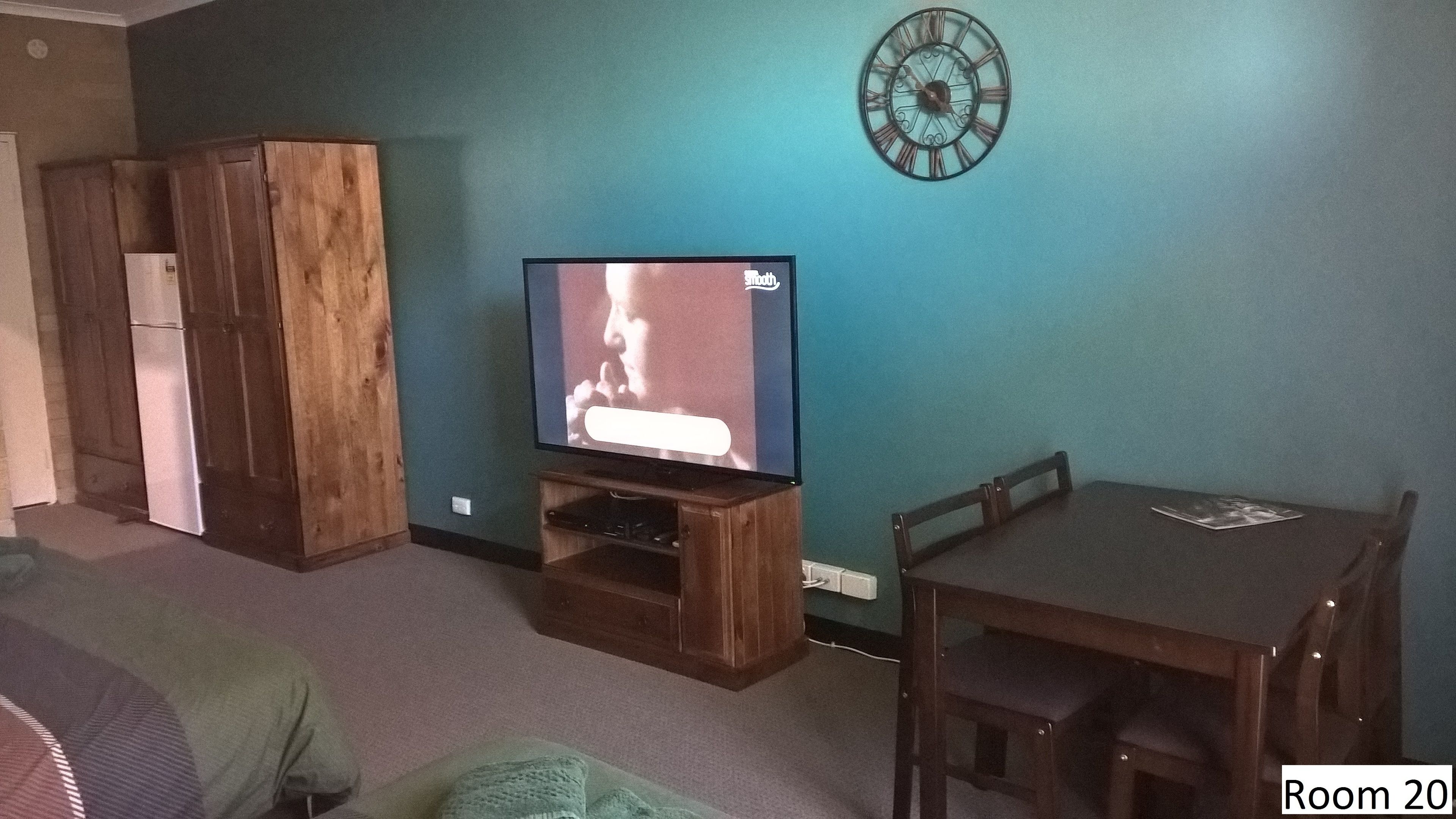 A room with teal walls, a wooden cabinet, a TV on a wooden stand, a dartboard, a dining table with chairs, and a refrigerator.