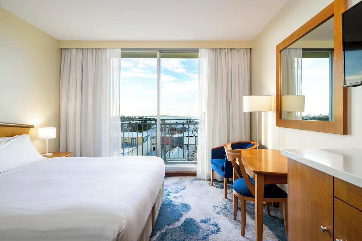 A hotel room with a large bed, a desk with two chairs, a TV, and a balcony with curtains.