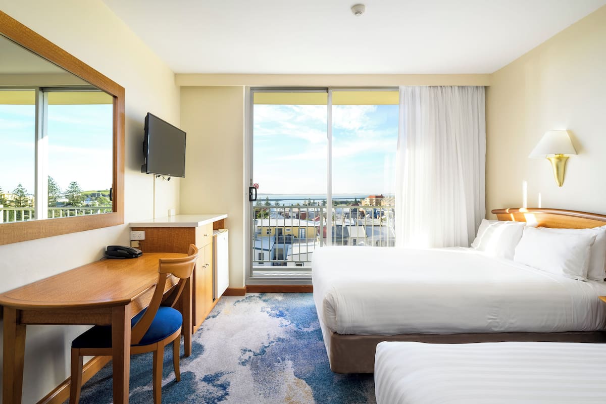 A hotel room with a bed, a desk with a chair, a TV, a lamp, and a view of the city through large windows.