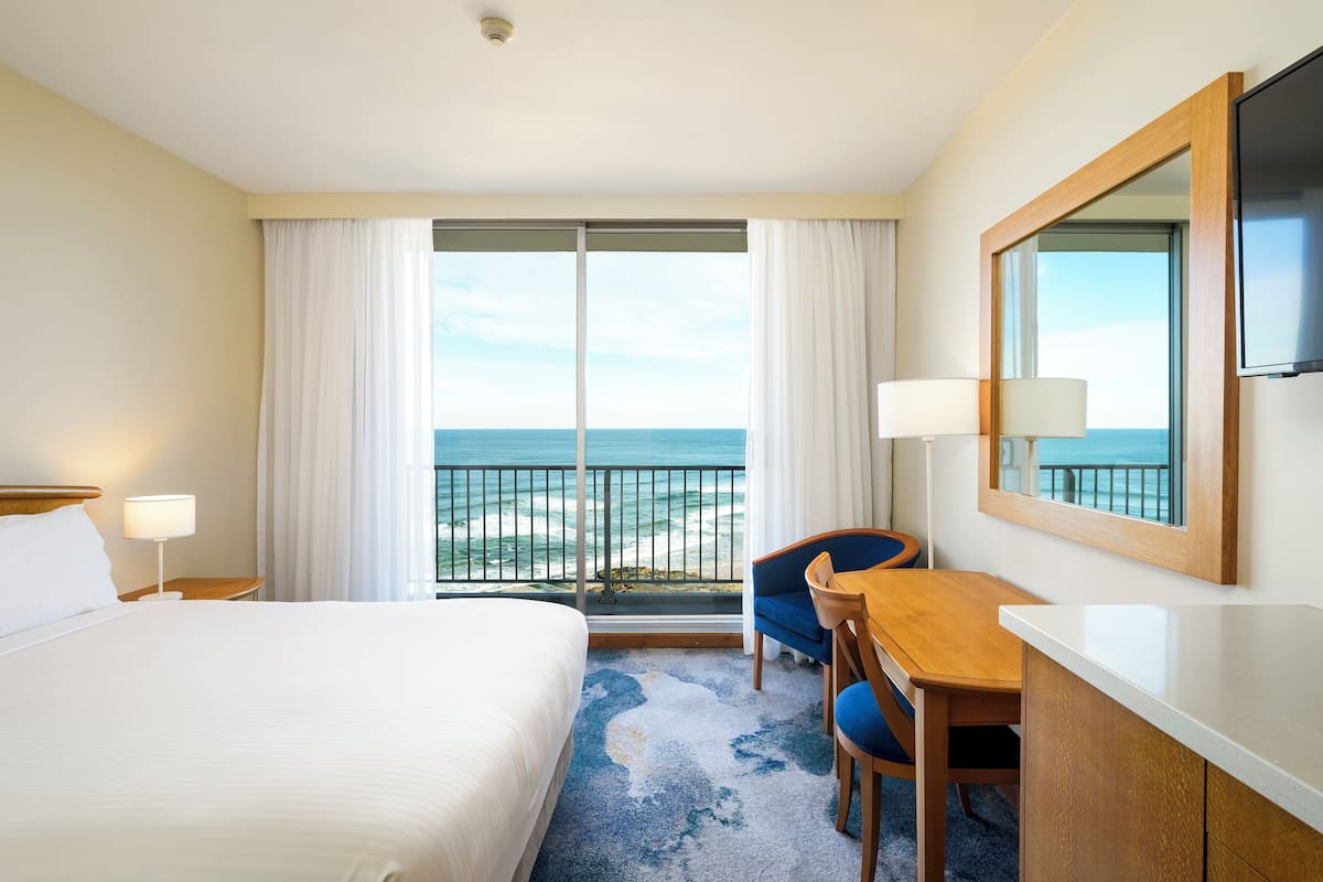 A hotel room with a bed, a desk, a chair, a TV, and a large window with a view of the ocean.