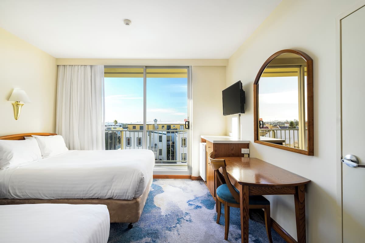 A hotel room with two beds, a desk, a chair, a television, and a large window offering a city view.