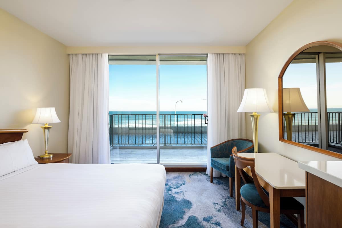A hotel room with a bed, a desk, a chair, a large mirror, and a balcony with a view of the ocean.