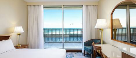 A hotel room with a bed, a desk, a chair, a large mirror, and a balcony with a view of the ocean.