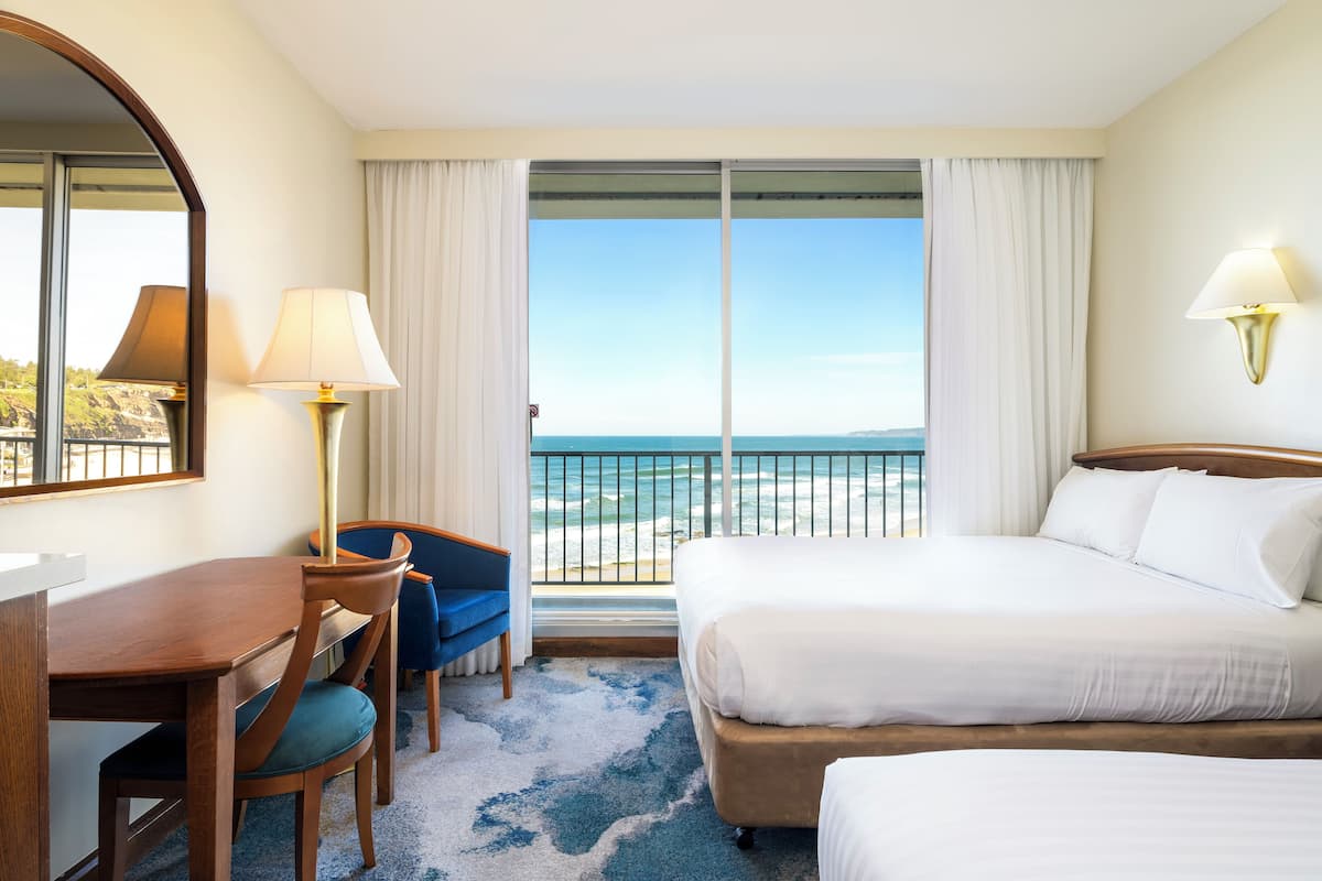 A hotel room with a large bed, a desk, a chair, and a view of the ocean through a sliding door.