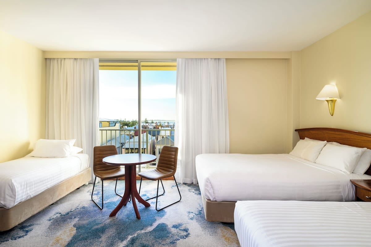 A hotel room with two beds, a round table, and a chair. There is a balcony with a view of buildings and a railing.