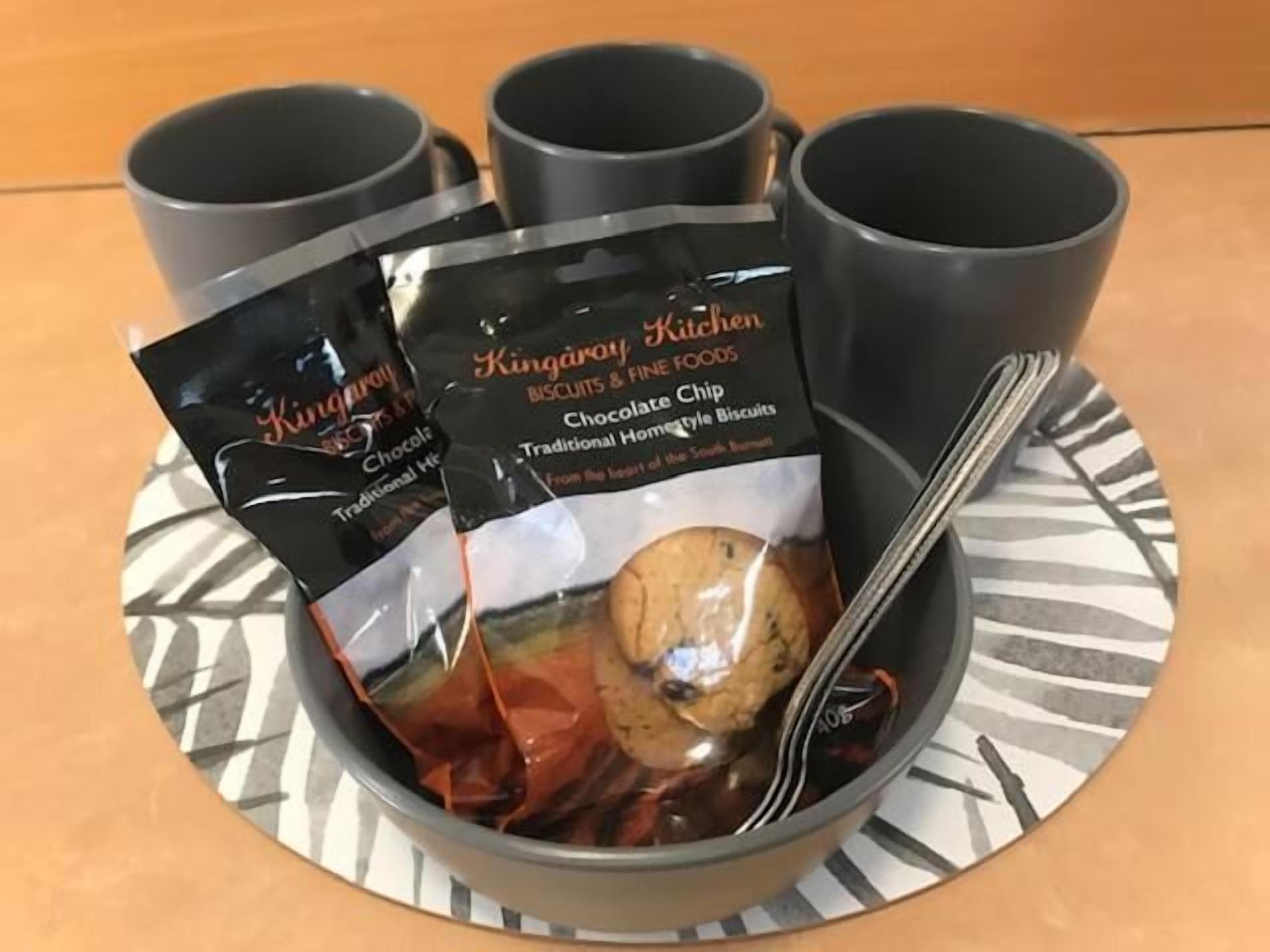 Una bandeja con tres tazas negras y un cuenco que contiene una bolsa de galletas con chispas de chocolate y una cuchara.