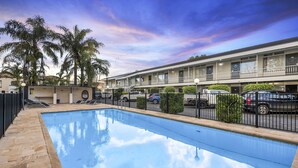 A swimming pool with a tiled deck, a multi-story building with balconies, and parked cars. - Park Motor Inn (East Toowoomba)