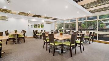 A spacious conference room with multiple tables and chairs, large windows offering a view of the outdoors, and a modern ceiling design.