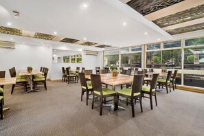 A spacious conference room with multiple tables and chairs, large windows offering a view of the outdoors, and a modern ceiling design.