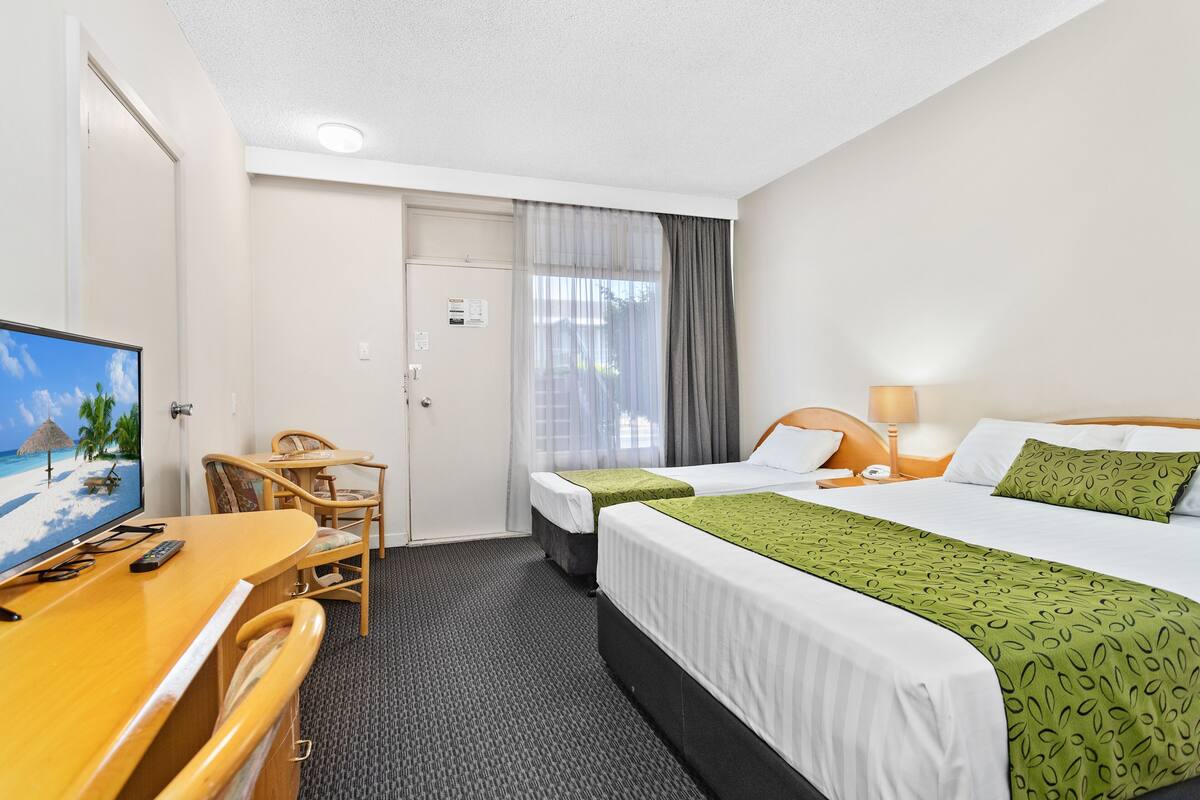 Quarto de hotel com escrivaninha, duas camas, televisão e janela com cortinas.