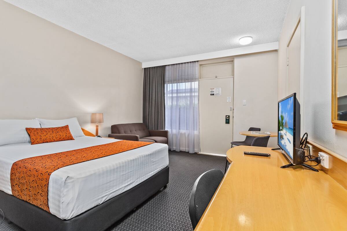 Quarto de hotel com cama, escrivaninha, cadeira, televisão e porta.