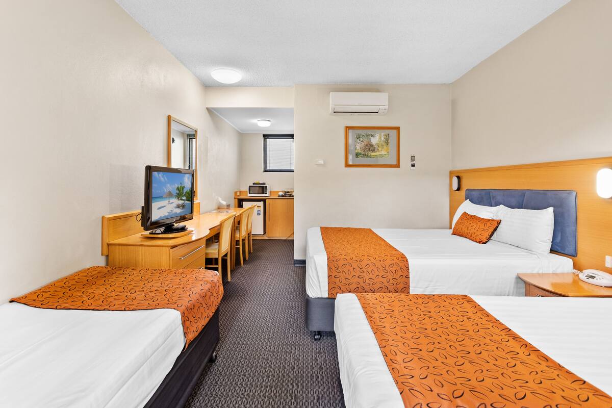Quarto de hotel com duas camas, uma escrivaninha, televisão e uma pequena cozinha.