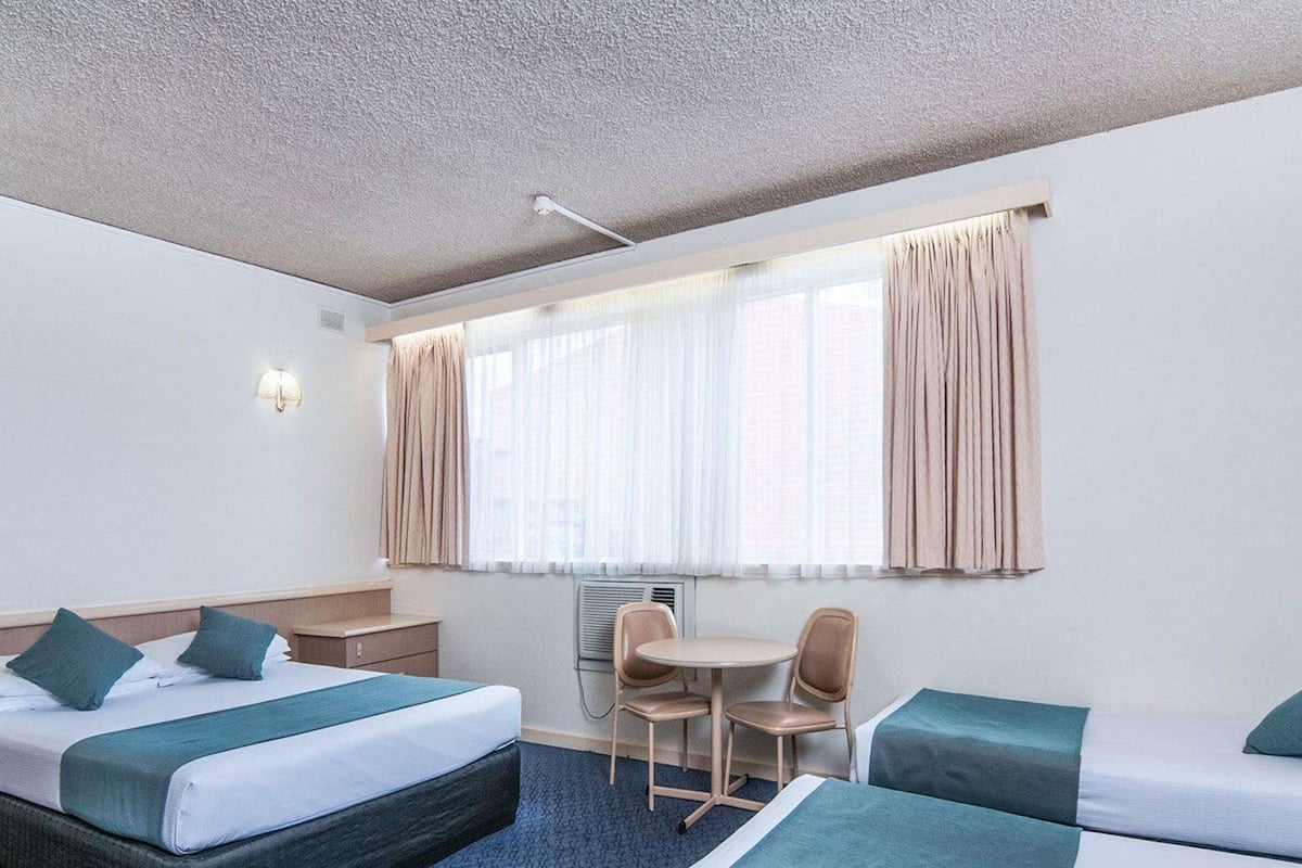 Hotellihuone, jossa on kaksi sänkyä, pieni pöytä, tuoli ja ikkuna, jossa on verhot.