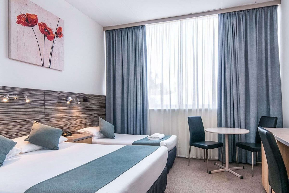 Hotellihuoneessa on kaksi sänkyä, työpöytä ja tuoli. Seinällä on taulu ja ikkunassa verhot.